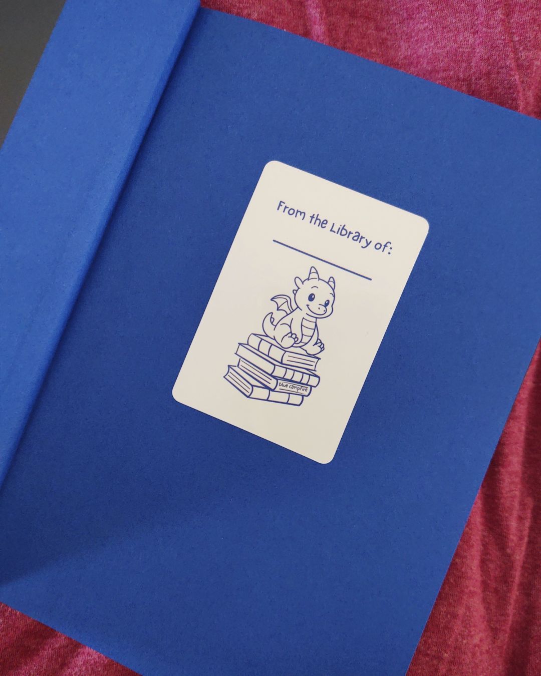 From the Library Of sticker with tiny blue dragon sitting atop a stack of books placed on a blue page in a book