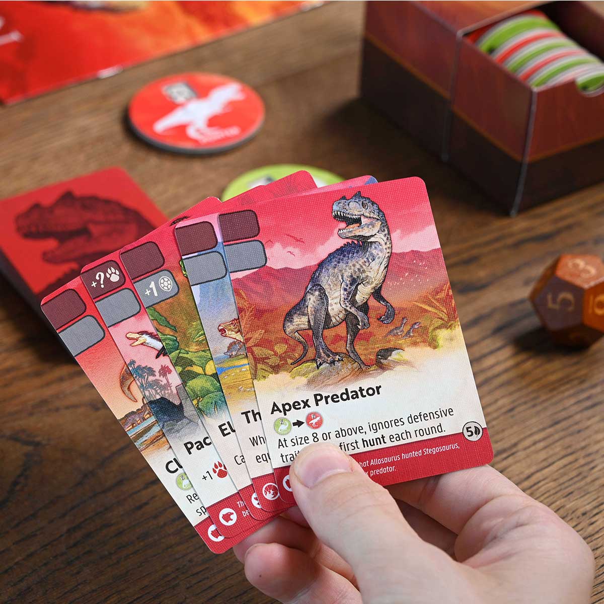 Hand holding a card with a dinosaur illustration labeled 'Apex Predator' on a wooden surface with game components.