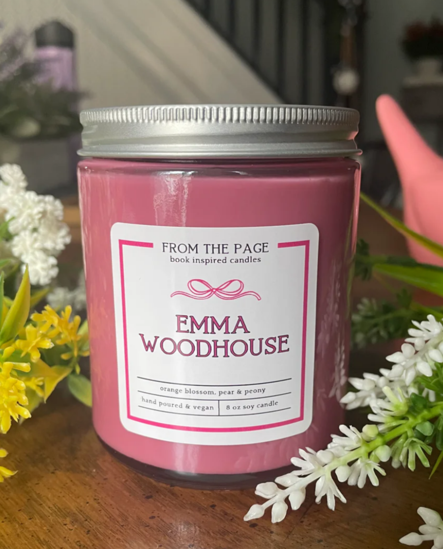 Pink candle labeled 'Emma Woodhouse' with a silver lid on a wooden surface surrounded by flowers.