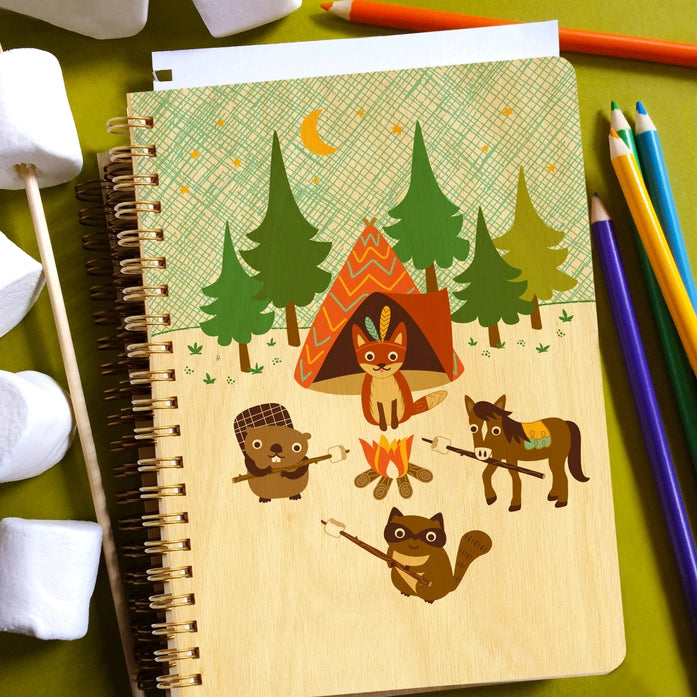 Notebook with forest-themed design featuring animals and a tent on a green surface with stationery items.