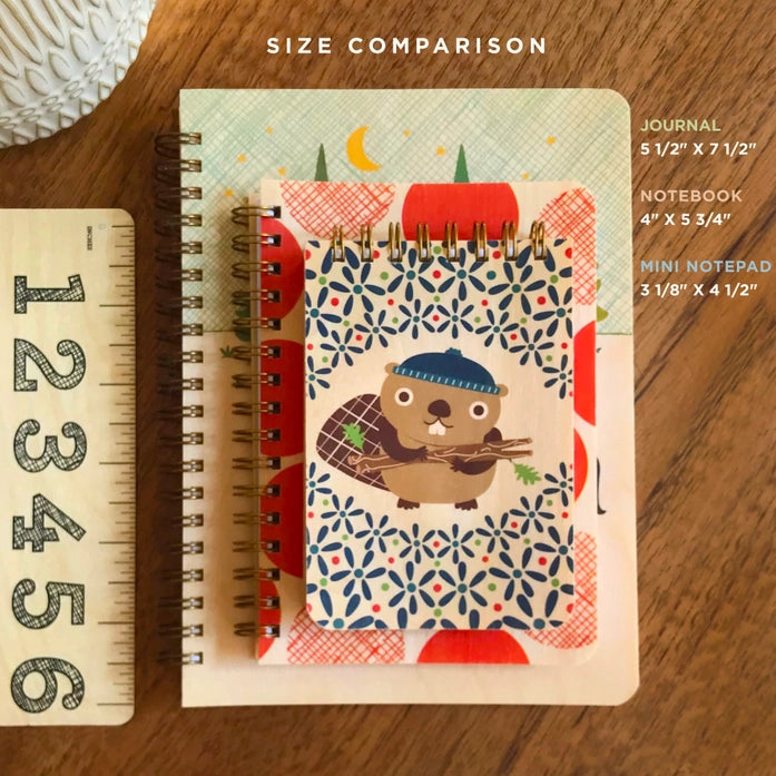 Size comparison of a journal, notebook, and mini notepad with a ruler on a wooden surface.